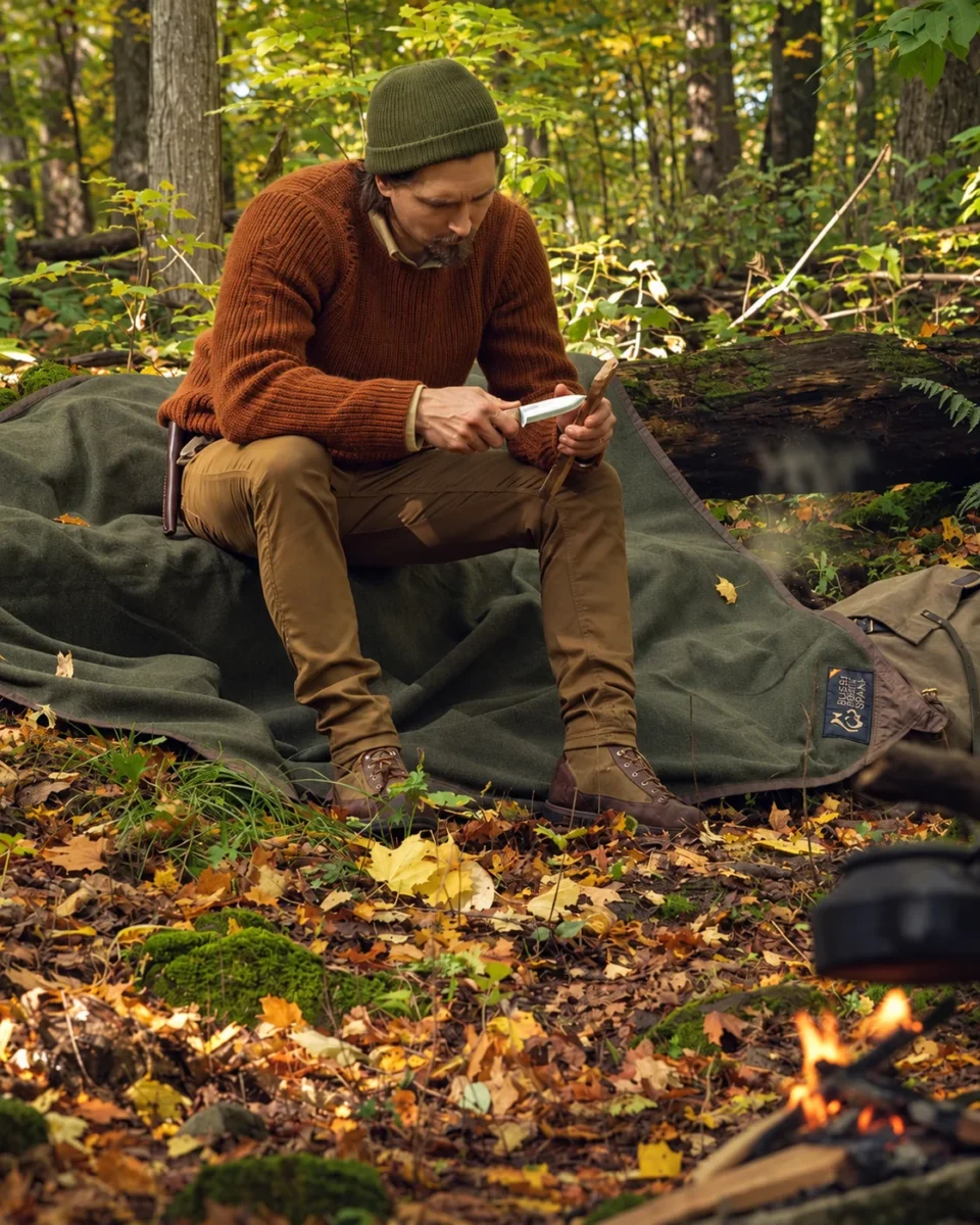 Bushcraft camper carving wood beside a campfire while sitting on an Oilskin and Wool Ground Cloth