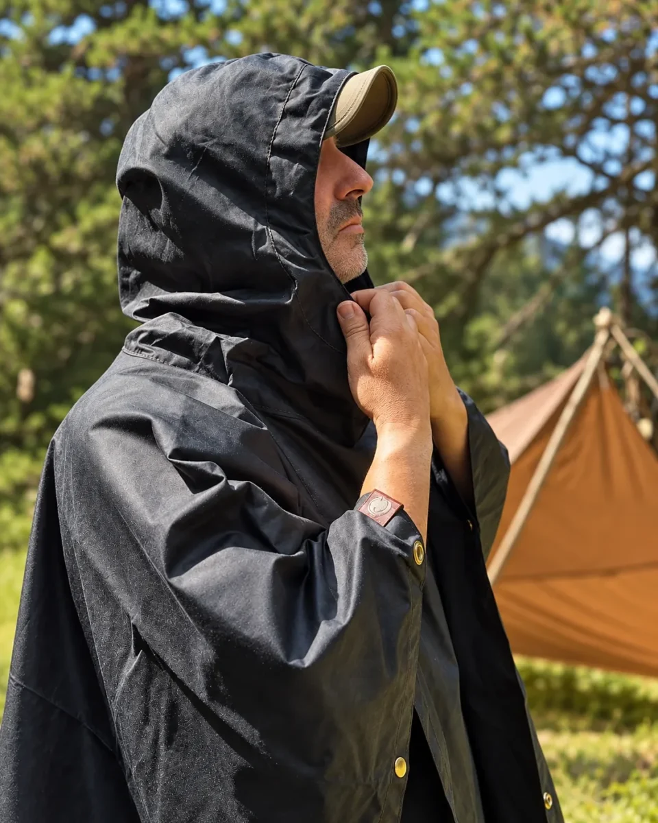 Black Oilskin Poncho Shelter with hood raised beside a woodland shelter